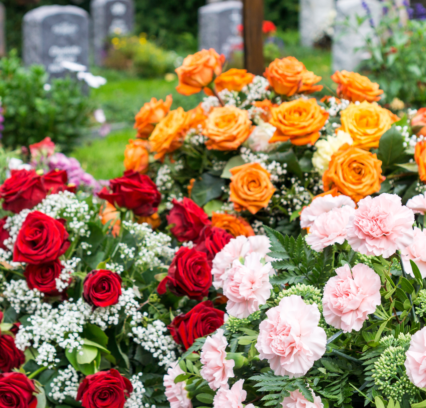 Bunte Blumenarrangements mit roten, orangefarbenen und rosa Rosen, weißen Blumen und grünen Blättern im Vordergrund eines Friedhofs mit Grabsteinen im Hintergrund.